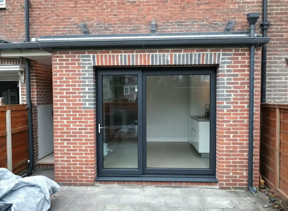 Een nieuwe antraciet schuifpui, net geplaatst in een verbrede opening in de bakstenen achtergevel, met een rollaag erboven en een afdekzeil op het terras
